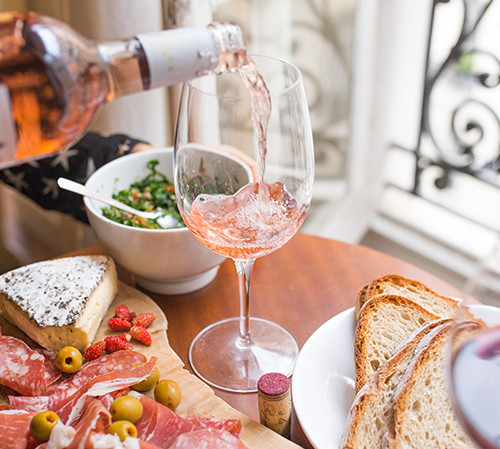 Wooden board with cold cuts & cheese next to glass of wine being poured